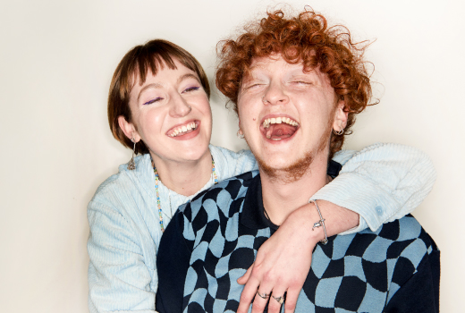 Zwei Personen, posieren vor einem neutralen Hintergrund. Eine Person hat den Arm um die Schulter der anderen gelegt.